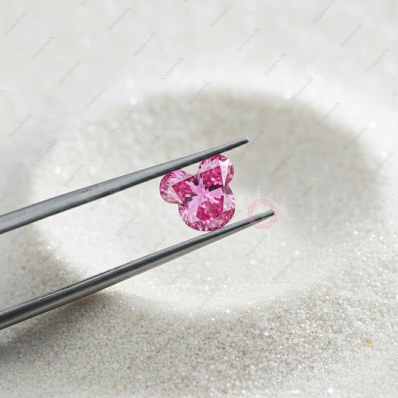 May include: A pink gemstone in the shape of a mouse head is held by silver tweezers. The gem is multi-faceted, reflecting light. The background is a white, textured surface, possibly sand or a similar material.