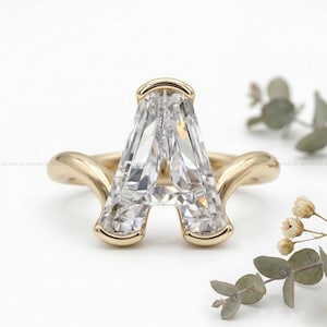 Könnte beinhalten: Ein goldener Ring mit einem großen, klaren, smaragdgeschliffenen Edelstein in Form des Buchstabens "A". Der Ring hat ein glattes, poliertes Goldband, das sich um den Edelstein legt. Der Ring ist vor einem weißen Hintergrund mit etwas Grün platziert.