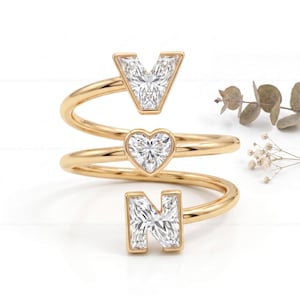Puede incluir: Anillo dorado con las letras "V", un corazón y "N", engastadas con piedras claras y brillantes. El anillo tiene un diseño en espiral que se enrolla alrededor del dedo. Fondo blanco.