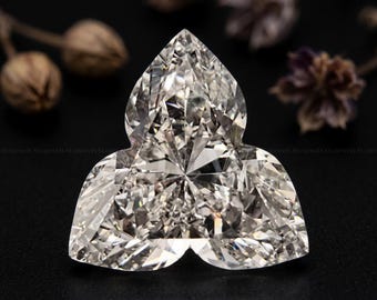 May include: A clear, colorless, faceted gemstone cut into a three-lobed, clover-like shape. The stone features numerous triangular and kite-shaped facets that reflect light, creating a brilliant, sparkling appearance against a dark background.