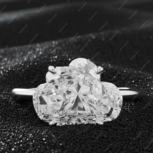 Op de afbeelding: Een zilveren ring met een grote, heldere, hartvormige edelsteen. De ring staat tegen een donkere, getextureerde achtergrond. De edelsteen heeft vele facetten die het licht reflecteren. De ring is een sieraad.