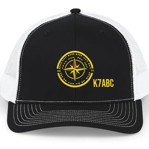 May include: A black and white trucker hat featuring a yellow embroidered compass design with the text K7ABC, CQ, and 73. The cap has a solid black front panel, a curved brim with black stitching, and white mesh side and back panels for ventilation.
