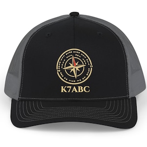 May include: A black and grey trucker cap featuring a gold compass graphic with a red needle tip and the text K7ABC printed in gold. The cap has a solid black front panel, a curved brim, and grey mesh side and back panels for ventilation.