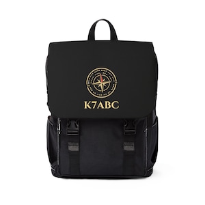 May include: A black backpack featuring a top flap with a gold compass graphic and the text K7ABC printed in gold. The bag includes a top handle, two side pockets, and two front straps with black plastic buckle closures for securing the main compartment.