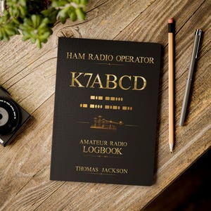 May include: A black logbook with gold text reading HAM RADIO OPERATOR, K7ABCD, AMATEUR RADIO LOGBOOK, and THOMAS JACKSON. The cover features gold Morse code symbols and a graphic of a telegraph key, positioned next to two pens.