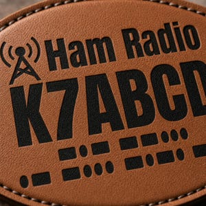 Puede incluir: Un parche de cuero color canela de forma ovalada con bordes cosidos que presenta texto negro en relieve que dice Ham Radio K7ABCD. El diseño incluye un icono de torre de radio negra a la izquierda y una serie de puntos y rayas en código Morse.