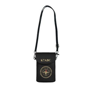 May include: A black crossbody bag with a long adjustable strap and a flap closure. The front features the gold text K7ABC above a circular gold compass graphic with a red needle accent. The bag has a smooth finish and silver-toned metal hardware.
