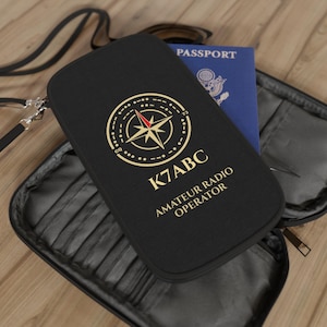 May include: A black travel organiser case with a detachable strap, featuring a gold compass graphic, the text K7ABC, and AMATEUR RADIO OPERATOR. A blue United States passport is tucked inside the open interior, which has multiple card slots.