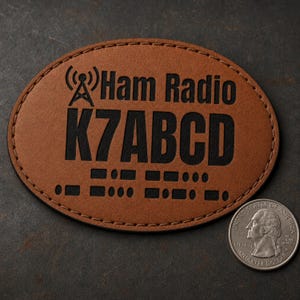 Op de afbeelding: Een ovale bruine leren patch met zwarte stiksels langs de rand. Het bevat een icoon van een radiotoren, de tekst "Ham Radio K7ABCD" en morsecode-symbolen. Een munt is ernaast geplaatst om de schaal van de patch aan te geven.