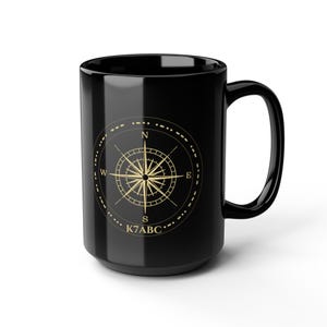 May include: A glossy black ceramic mug featuring a gold-colored compass rose graphic on the front. The compass design includes the cardinal directions N, S, E, and W, a circular border with Morse code-style dots and dashes, and the text K7ABC.
