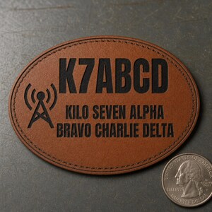 Puede incluir: Un parche ovalado de cuero marrón con el texto negro K7ABCD, las palabras KILO SEVEN ALPHA BRAVO CHARLIE DELTA y un icono de torre de radio. Una moneda estadounidense está colocada junto al parche para proporcionar una escala de tamaño.