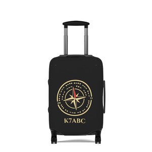 May include: A black rolling suitcase with a retractable handle and four wheels. The front features a gold compass rose graphic with a red needle tip, surrounded by circular markings, the letters CQ, the number 73, and the text K7ABC below it.