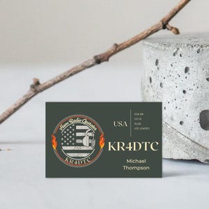 Puede incluir: Una tarjeta de visita con un logotipo de operador de radioaficionado, con la bandera estadounidense, llamas y el texto "KR4DTC". La tarjeta también incluye el nombre "Michael Thompson" y detalles de EE. UU. Una ramita y un bloque de hormigón en el fondo.
