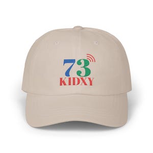 May include: A beige baseball cap featuring the number "73" in blue, a green "73" with a radio wave design, and the word "KIDXY" in red. The cap has a curved peak and a rounded crown.
