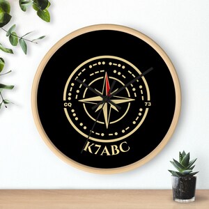 May include: A round wall clock with a light wood frame and a black face. The center features a gold compass rose design with a red-tipped needle, surrounded by Morse code patterns, the letters CQ, the number 73, and the call sign K7ABC in gold.
