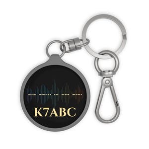 Peut inclure: Un porte-clés rond en métal avec un insert circulaire noir orné d'un graphique d'onde sonore coloré, d'une ligne de code Morse et du texte K7ABC en lettres dorées. Le porte-clés comprend un anneau fendu argenté et un mousqueton pivotant.