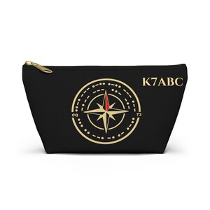 May include: A black zippered pouch featuring a gold-colored compass rose graphic with a red accent on the top needle. The design includes Morse code-style dots and dashes around the compass, the letters CQ and 73, and the text K7ABC in gold.