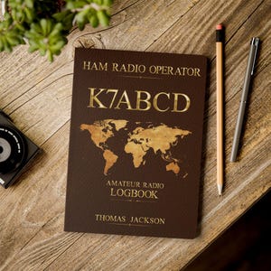 May include: A dark brown ham radio operator logbook featuring a gold world map graphic, the text HAM RADIO OPERATOR, K7ABCD, AMATEUR RADIO LOGBOOK, and THOMAS JACKSON. The cover has a textured finish with gold lettering and map details.