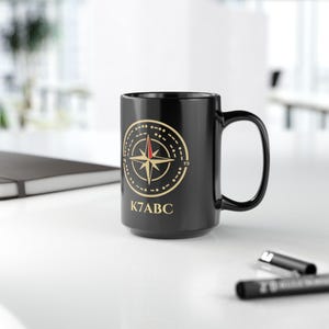 May include: A black ceramic mug featuring a gold circular compass design with a red needle accent. The compass is surrounded by a dotted ring pattern and the text K7ABC is printed in gold lettering below the graphic on the front of the mug.