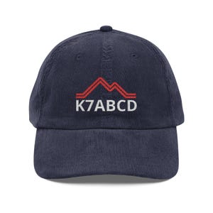 Könnte beinhalten: Eine marineblaue Baseballkappe aus Kord mit einer Stickerei auf der Vorderseite. Das Design besteht aus einer roten Bergumrissgrafik über dem weißen gestickten Text K7ABCD, der mittig auf der Vorderseite der Kappe platziert ist.