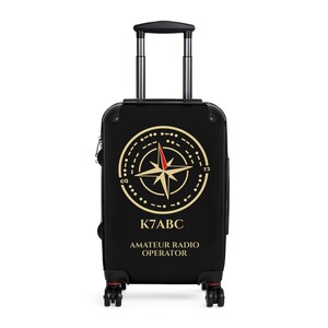 May include: A black carry-on suitcase with a retractable handle and four wheels. The front features a gold compass rose graphic with a red needle, Morse code symbols, the text CQ, 73, K7ABC, and AMATEUR RADIO OPERATOR in gold lettering.