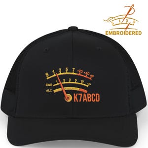 May include: A black baseball cap featuring an embroidered radio signal meter graphic on the front. The design includes yellow and orange scales labeled S, SWR, and ALC, with the text K7ABCD in orange. The cap has a structured crown and mesh back.