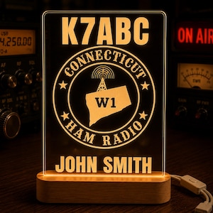 May include: A rectangular acrylic LED sign with a wooden base, featuring the text K7ABC, CONNECTICUT, W1, HAM RADIO, and JOHN SMITH. The design includes a graphic of a radio tower and the state of Connecticut, all illuminated in a warm amber glow.
