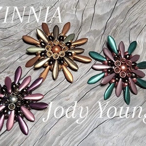 May include: Three Zinnia flower brooches in bronze, green, and purple. Each brooch has a central cluster of beads surrounded by petal-shaped elements. The brooches are displayed on a wooden surface, with the name "Jody Young" visible.
