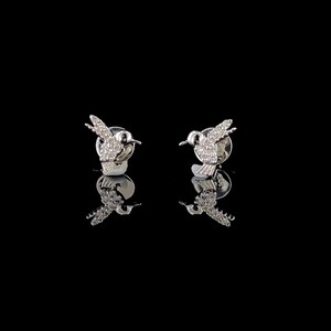 Peut inclure: Une paire de boucles d'oreilles clous en forme de colibri argentés. Chaque boucle d'oreille présente un motif d'oiseau détaillé avec des ailes déployées, incrustées de petites pierres scintillantes. Les boucles d'oreilles sont posées sur un fond noir, avec leurs reflets visibles en dessous.