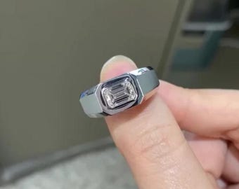 May include: A silver ring with a rectangular, emerald-cut gemstone. The ring has a modern, geometric design with a polished finish. The gemstone is clear and appears to be set in a bezel setting.