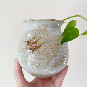 May include: A white ceramic vase with brown and gray glaze. The vase has a rounded shape and a wide opening. A green plant is partially visible in the opening.