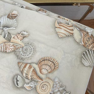 May include: A cream-colored fabric featuring a seashell pattern in shades of brown, beige, and gray. The design includes various seashell shapes and sizes, creating a coastal or beach-themed aesthetic. The fabric is folded over a gold-framed glass table.