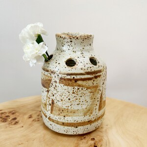 May include: A white ceramic vase with brown speckles and geometric designs. The vase has several holes around the top for flower stems. White carnations are in the vase.