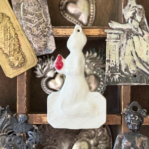 May include: A white translucent figurine of a kneeling figure holding a small red heart, positioned centrally among various metallic wall plaques featuring religious and figurative motifs, including hearts and human forms, in a wooden grid.