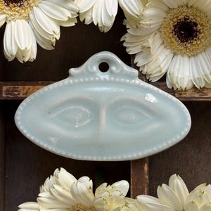 May include: A pale blue ceramic decorative tray with a face design, surrounded by white gerbera daisies. The tray has a small hole at the top and a beaded edge. The background is a dark wooden surface.