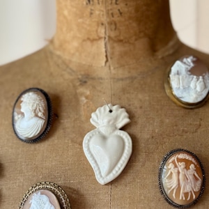 Könnte beinhalten: Eine Sammlung antiker Cameo-Broschen und -Anhänger. Die Broschen zeigen detaillierte Reliefschnitzereien klassischer Figuren und Szenen, gefasst in verzierten Metallrahmen. Ein weißer herzförmiger Keramikanhänger ist ebenfalls zu sehen.
