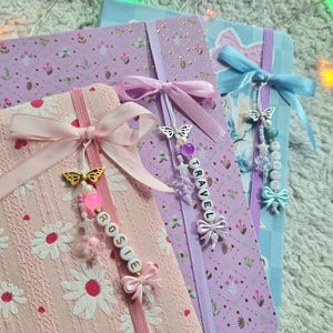 Op de afbeelding: Drie decoratieve tijdschriften met bloemen- en strikdesigns. Eén tijdschrift is roze met witte madeliefjes en de naam "Rosie" op een kralenbedel. Een andere is paars met het woord "TRAVEL" op een bedel. De derde is blauw met een strik.