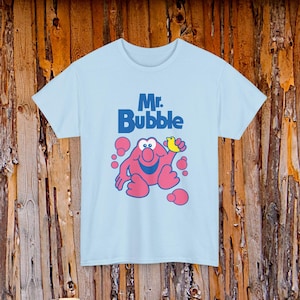 May include: Light blue t-shirt featuring the text "Mr. Bubble" in blue, with a cartoon character holding a yellow duck. The character is pink with blue eyes and pink bubbles around it. The shirt has short sleeves and a crew neck.
