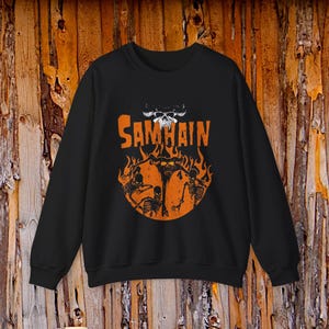 May include: A black crewneck sweatshirt featuring a graphic print in orange and white. The design displays the text SAMHAIN above a circular orange flame motif containing three black skeleton figures and a horned skull icon above the text.