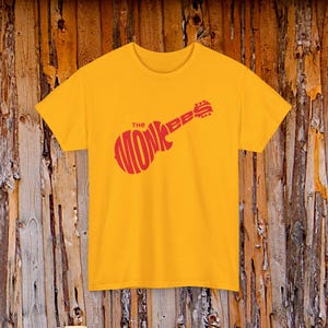 Może przedstawiać: Jaskrawożółty T-shirt z krótkim rękawem z czerwonym nadrukiem graficznym na klatce piersiowej. Grafika ma kształt gitary i zawiera napis "THE MONKEES" stylizowaną czcionką, przy czym słowo "THE" znajduje się nad korpusem gitary.