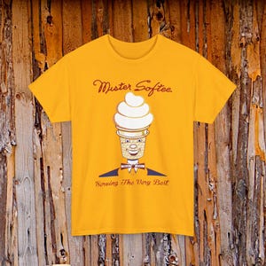Puede incluir: Una camiseta de color amarillo mostaza con un gráfico del personaje de cono de helado Mister Softee. El diseño incluye el texto Mister Softee en la parte superior y Serving The Very Best en la inferior, con el personaje usando pajarita y chaqueta.