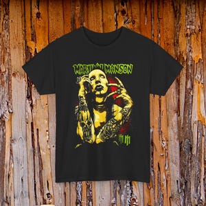 Może przedstawiać: Czarny t-shirt z żółto-czerwoną grafiką przedstawiającą wytatuowaną osobę trzymającą papierosa. Tekst "Marilyn Manson" jest wydrukowany na zielono u góry. Koszulka ma okrągły dekolt i krótkie rękawy.