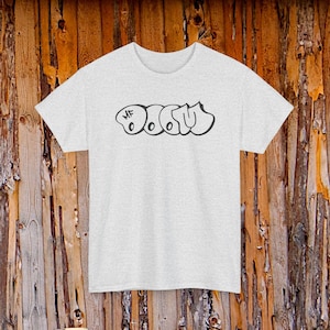 Könnte beinhalten: Hellgraues T-Shirt mit dem schwarzen Graffiti-Schriftzug "MF DOOM". Das Shirt hat einen Rundhalsausschnitt und kurze Ärmel. Der Hintergrund ist eine Holzwand.