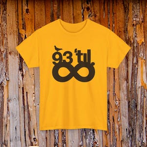 Puede incluir: Camiseta de color amarillo mostaza con un diseño gráfico negro. El diseño incluye el texto "93'tıl" encima de un símbolo de infinito. Un pequeño pájaro está posado encima del texto. La camiseta es de material suave.