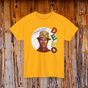 May include: Mustard yellow t-shirt featuring a portrait of a man wearing a hat, inside a white circle. The text "Are We Not Men?" is above the portrait, and the band name "DEVO" is printed in colorful letters to the right.