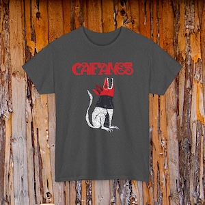 Può includere: Una t-shirt a maniche corte grigio scuro con la scritta rossa CAIFANES sopra la grafica di un animale bianco stilizzato con testa rossa e collare nero. L'animale è raffigurato in posizione seduta con la testa rivolta verso l'alto e la bocca aperta.