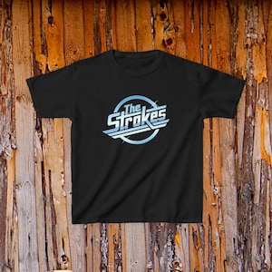 May include: A black short-sleeved t-shirt featuring a centered graphic of the band name The Strokes in a light blue and white stylized font enclosed within a circular border with subtle starburst accents on the lettering and the circle.