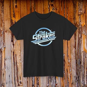 May include: Black t-shirt featuring the band name "The Strokes" in a light blue and white design. The text is inside a light blue circle with white lines. The shirt is displayed against a wooden background.