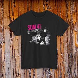 May include: A black short-sleeve t-shirt featuring a graphic print with the pink text SUM 41 and the white text UNDERCLASS HERO on a banner. The graphic includes a black and white image of a person standing next to a fire extinguisher.