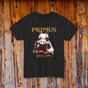 May include: Black t-shirt featuring the band name "PRIMUS" in gold lettering above a graphic of a pig's face. The words "Pork Soda" are written in gold script below the pig illustration.
